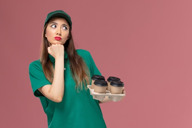 Vue de face femme courrier en uniforme vert et cape tenant des tasses de café de livraison posant et pensant sur la livraison uniforme de service de mur rose