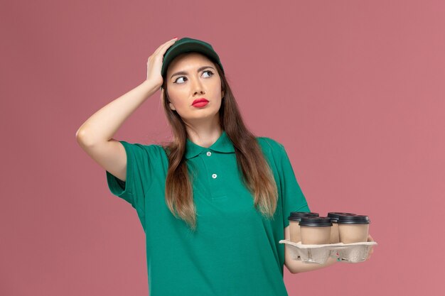 Vue de face femme courrier en uniforme vert et cape tenant des tasses de café de livraison et de la réflexion sur le travail de livraison uniforme de service de mur rose