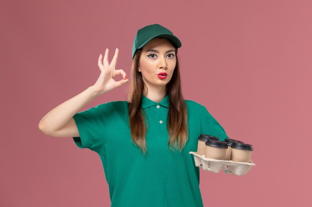 Vue de face femme courrier en uniforme vert et cape tenant des tasses de café de livraison sur le service de bureau rose clair travail de livraison uniforme