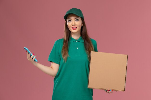 Vue de face femme courrier en uniforme vert tenant la boîte de livraison de nourriture à l'aide de téléphone sur le mur rose entreprise service de livraison uniforme de travail