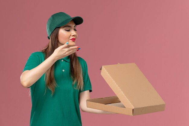 Vue de face femme courrier en uniforme vert tenant la boîte de livraison de nourriture sentant sur le mur rose service de l'entreprise livraison uniforme