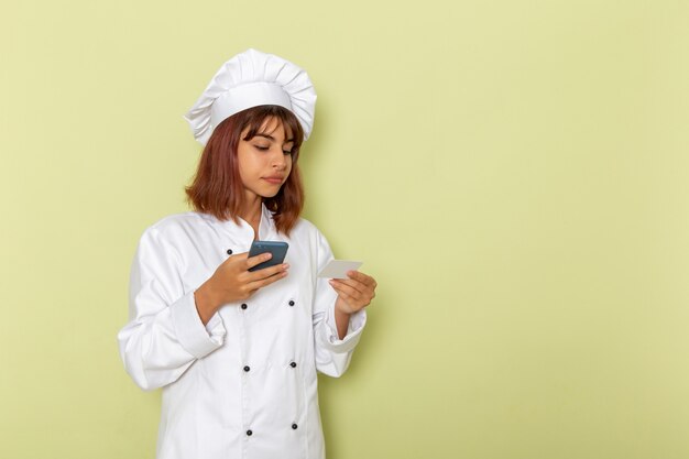 Vue de face femme cuisinier en costume de cuisinier blanc à l'aide de son smartphone sur une surface verte
