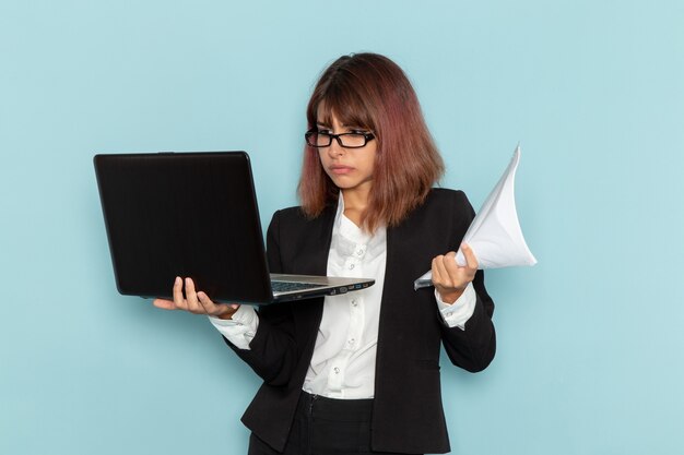 Vue de face femme employée de bureau en costume strict à l'aide d'un ordinateur portable et tenant des papiers sur une surface bleu clair