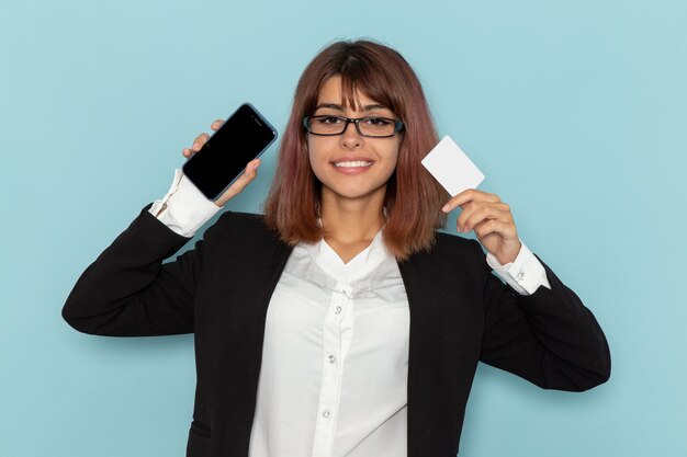 Vue de face femme employée de bureau en costume strict tenant une carte blanche et un téléphone sur une surface bleu clair