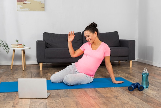 Vue de face de la femme enceinte à la maison exercice sur tapis avec ordinateur portable