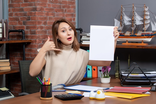 Vue de face d'une femme étonnée faisant appel moi signe de téléphone assis au bureau