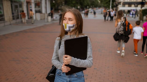 Vue de face femme avec masque médical tenant son ordinateur portable à l'extérieur