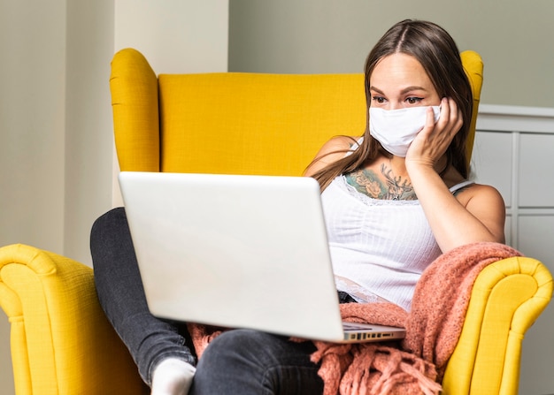 Vue de face de la femme avec un masque médical travaillant sur un ordinateur portable à partir d'un fauteuil pendant la pandémie