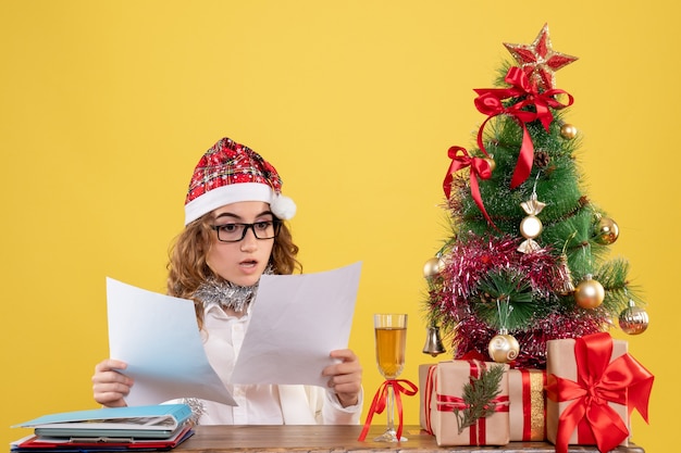 Vue de face femme médecin assis avec arbre de Noël présente et tenant des documents sur fond jaune