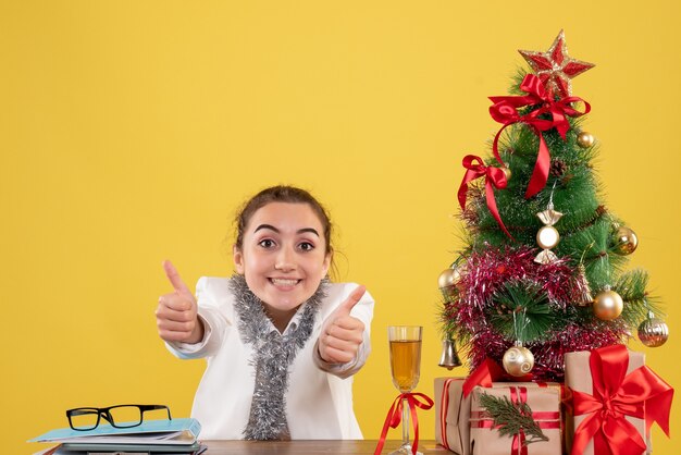 Vue de face femme médecin assis autour de cadeaux de Noël et arbre sur fond jaune