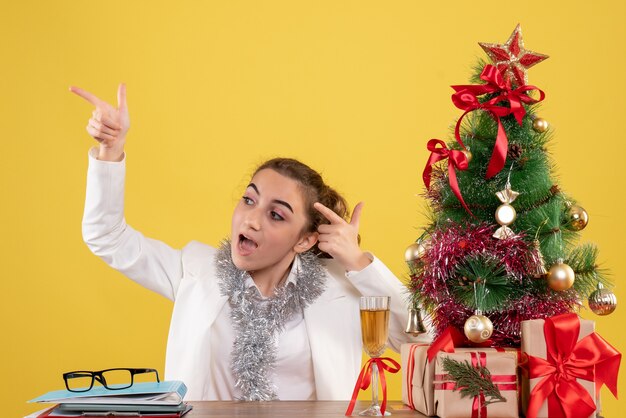 Vue de face femme médecin assis autour de cadeaux de Noël et arbre sur fond jaune