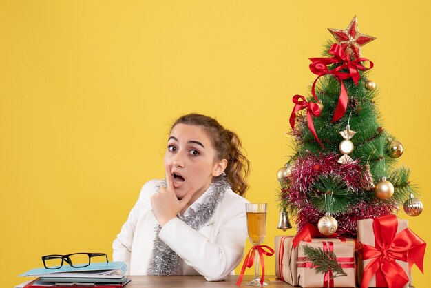 Vue de face femme médecin assis autour de cadeaux de Noël et arbre sur fond jaune