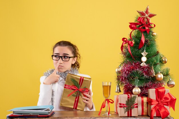 Vue de face femme médecin assis autour de cadeaux de Noël et arbre sur fond jaune