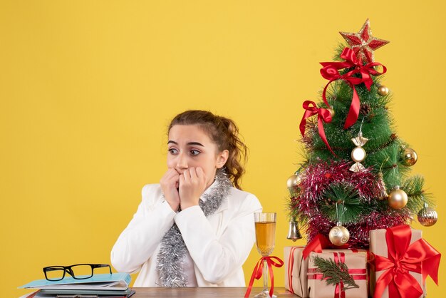 Vue de face femme médecin assis autour des cadeaux de Noël et arbre peur sur fond jaune