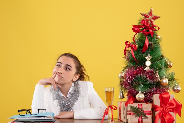 Vue de face femme médecin assis autour de cadeaux de Noël et arbre s'ennuie