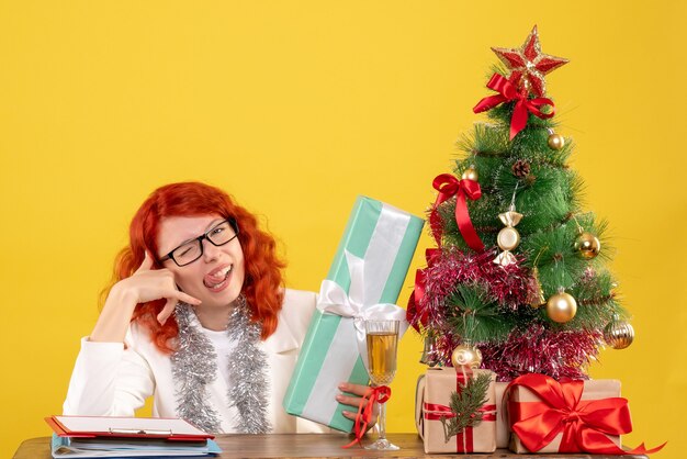 Vue de face femme médecin assis avec des cadeaux de Noël et arbre sur un bureau jaune