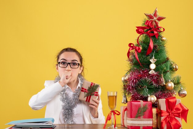 Vue de face femme médecin assis avec des cadeaux de Noël et arbre sur un bureau jaune