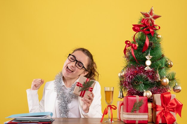 Vue de face femme médecin assis avec des cadeaux de Noël et arbre sur fond jaune