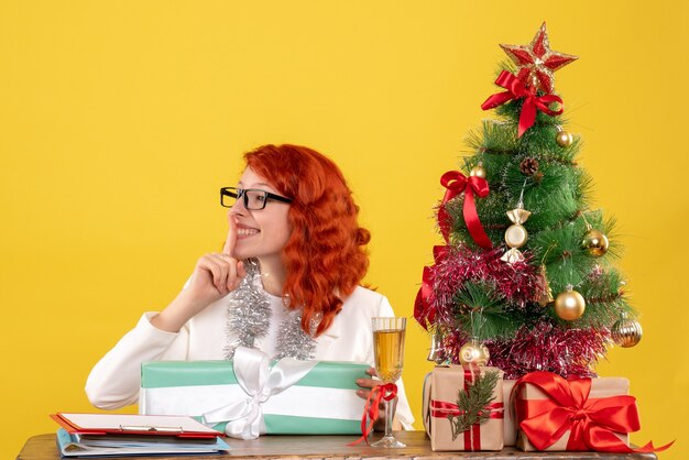 Vue de face femme médecin assis avec des cadeaux de Noël et arbre sur fond jaune