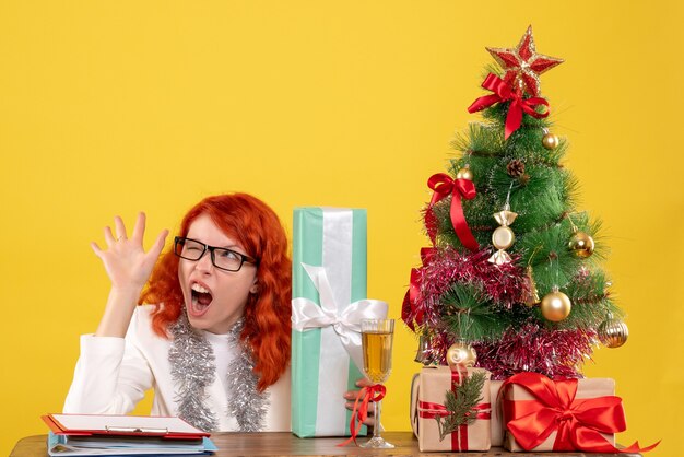 Vue de face femme médecin assis avec des cadeaux de Noël et arbre sur fond jaune