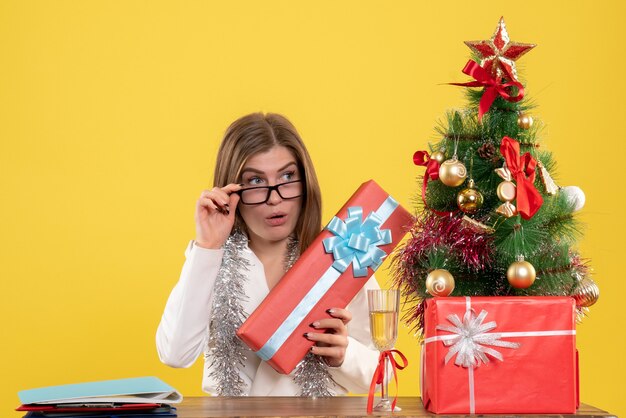 Vue de face femme médecin assis avec des cadeaux de Noël et arbre sur jaune