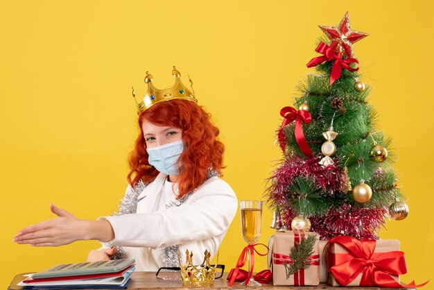 Vue de face femme médecin assis avec des cadeaux de Noël et portant une couronne sur un bureau jaune