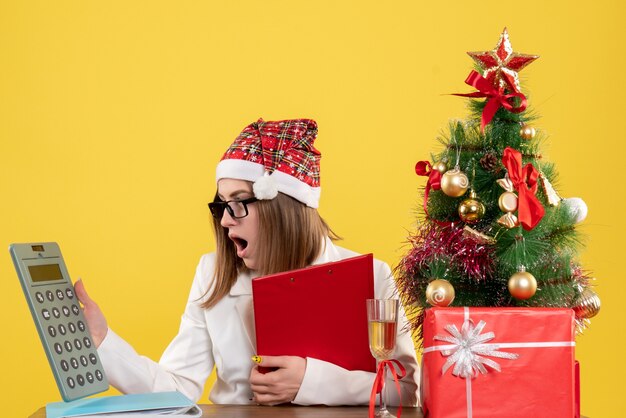 Vue de face femme médecin assis avec des cadeaux de Noël tenant calculatrice sur fond jaune