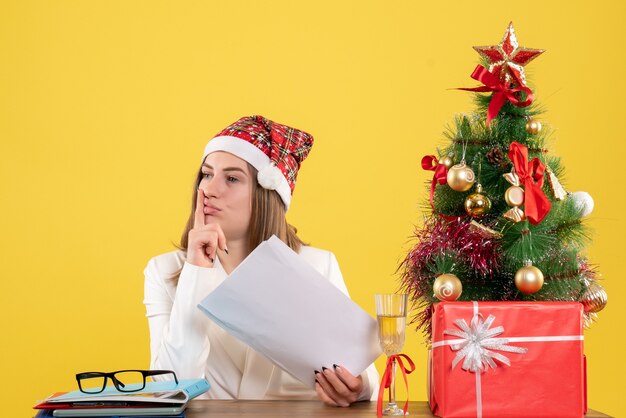 Vue de face femme médecin assis avec des cadeaux de Noël tenant des documents sur fond jaune