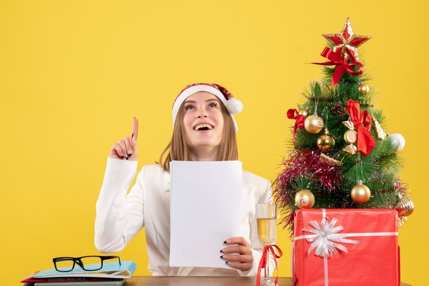 Vue de face femme médecin assis avec des cadeaux de Noël tenant des documents sur fond jaune