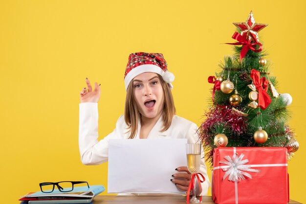 Vue de face femme médecin assis avec des cadeaux de Noël tenant des documents sur fond jaune