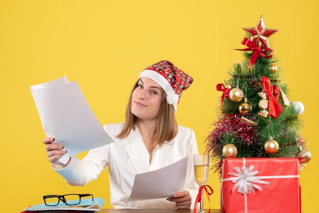 Vue de face femme médecin assis avec des cadeaux de Noël tenant des documents sur fond jaune