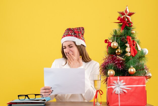 Vue de face femme médecin assis avec des cadeaux de Noël tenant des documents sur fond jaune