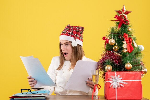 Vue de face femme médecin assis avec des cadeaux de Noël tenant des documents sur fond jaune