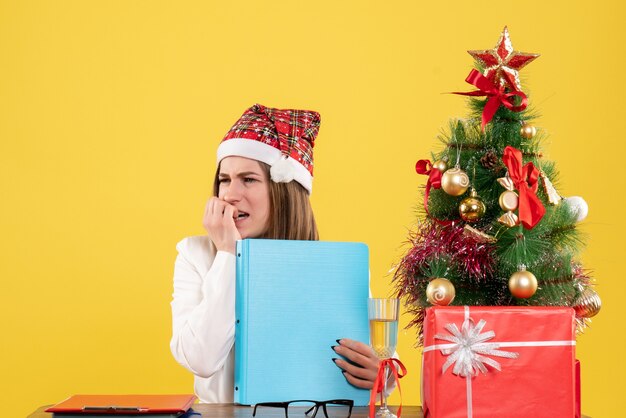 Vue de face femme médecin assis avec des cadeaux de Noël tenant des fichiers sur fond jaune