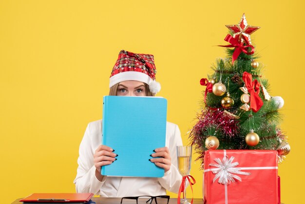 Vue de face femme médecin assis avec des cadeaux de Noël tenant des fichiers sur fond jaune