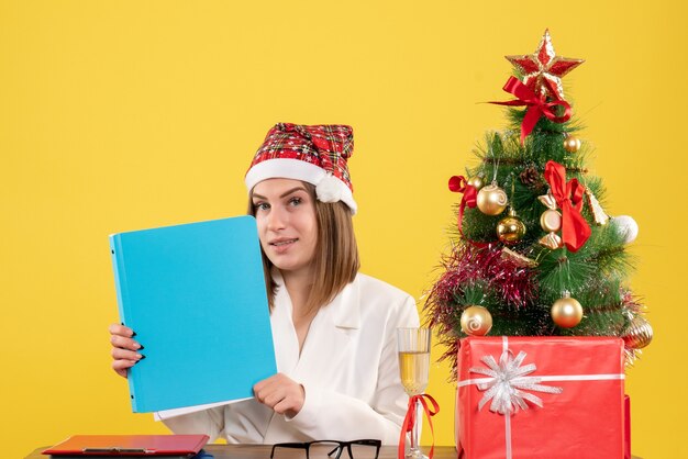 Vue de face femme médecin assis avec des cadeaux de Noël tenant des fichiers sur fond jaune