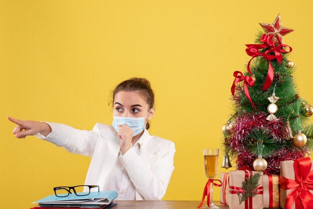 Vue de face femme médecin assis dans un masque de protection sur un bureau jaune avec arbre de Noël et coffrets cadeaux