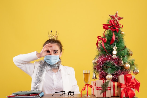 Vue de face femme médecin assis dans un masque stérile sur fond jaune avec arbre de Noël et coffrets cadeaux