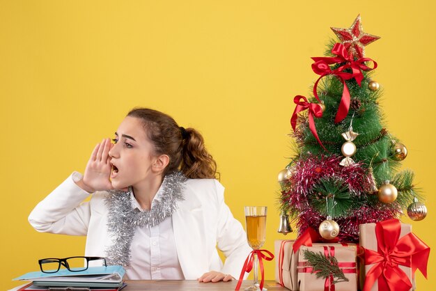 Vue de face femme médecin assis derrière sa table appelant quelqu'un sur fond jaune avec arbre de Noël et coffrets cadeaux