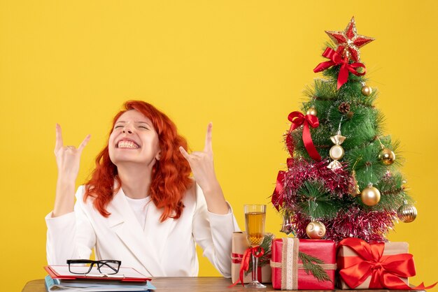 Vue de face femme médecin assis derrière sa table avec des cadeaux de Noël et arbre sur un bureau jaune