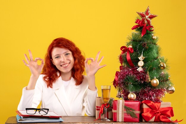 Vue de face femme médecin assis derrière sa table avec des cadeaux de Noël et arbre sur fond jaune