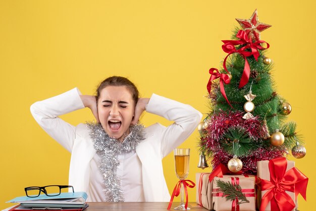 Vue de face femme médecin assis derrière sa table et fermant ses oreilles sur fond jaune avec arbre de Noël et coffrets cadeaux