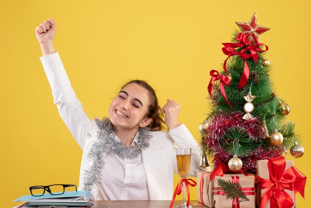 Vue de face femme médecin assis derrière sa table sur un fond jaune avec arbre de Noël et coffrets cadeaux