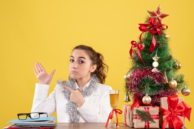 Vue de face femme médecin assis derrière sa table sur un fond jaune avec arbre de Noël et coffrets cadeaux