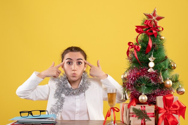 Vue de face femme médecin assis derrière sa table sur un fond jaune avec arbre de Noël et coffrets cadeaux