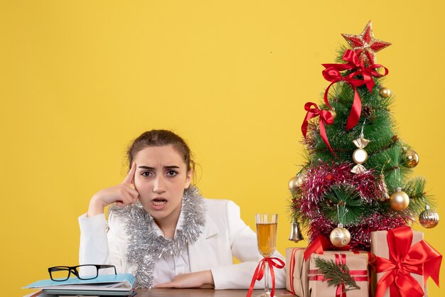 Vue de face femme médecin assis derrière sa table sur un fond jaune avec arbre de Noël et coffrets cadeaux