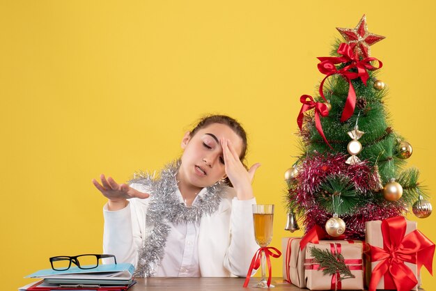 Vue de face femme médecin assis derrière sa table sur un fond jaune avec arbre de Noël et coffrets cadeaux