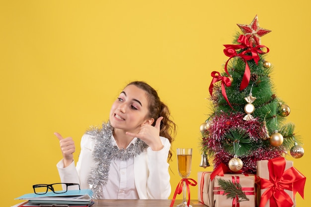 Vue de face femme médecin assis derrière sa table sur un fond jaune avec arbre de Noël et coffrets cadeaux