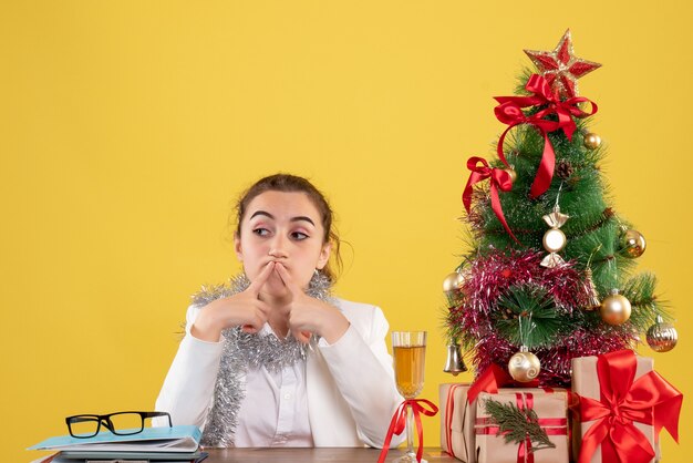 Vue de face femme médecin assis derrière sa table s'ennuie sur fond jaune avec arbre de Noël et coffrets cadeaux