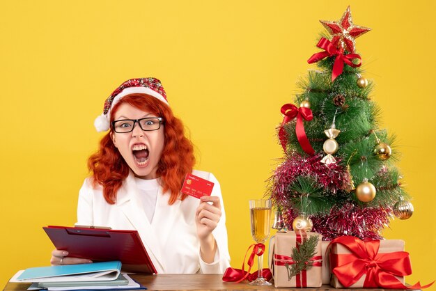Vue de face femme médecin assis derrière sa table et tenant une carte bancaire sur un bureau jaune avec arbre de Noël et coffrets cadeaux